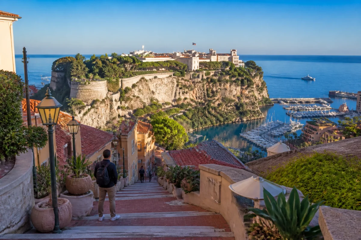 Vue panoramique de Monaco au matin, avec le palais princier, le port et un voyageur dominant la Côte d’Azur.